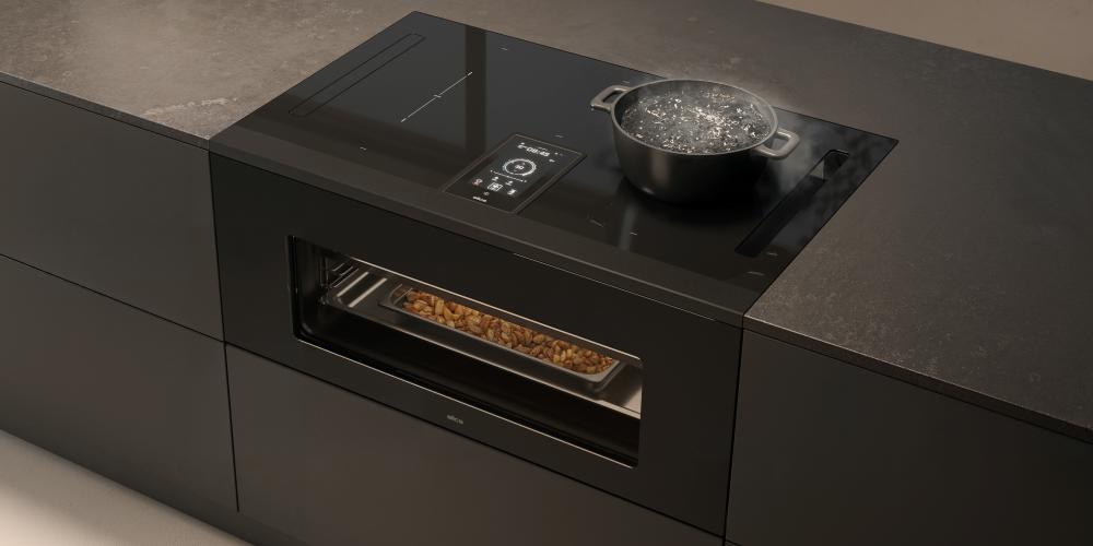 A built-in hob and oven combo on a black island. The hob has a pot with boiling water on it, while the oven is baking food on a tray.