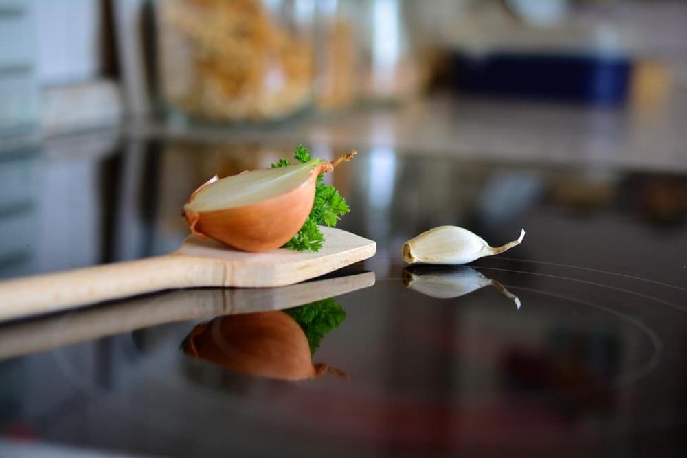 A wooden spatula, half an onion, a clove of garlic, and some herbs are placed on an induction hob.