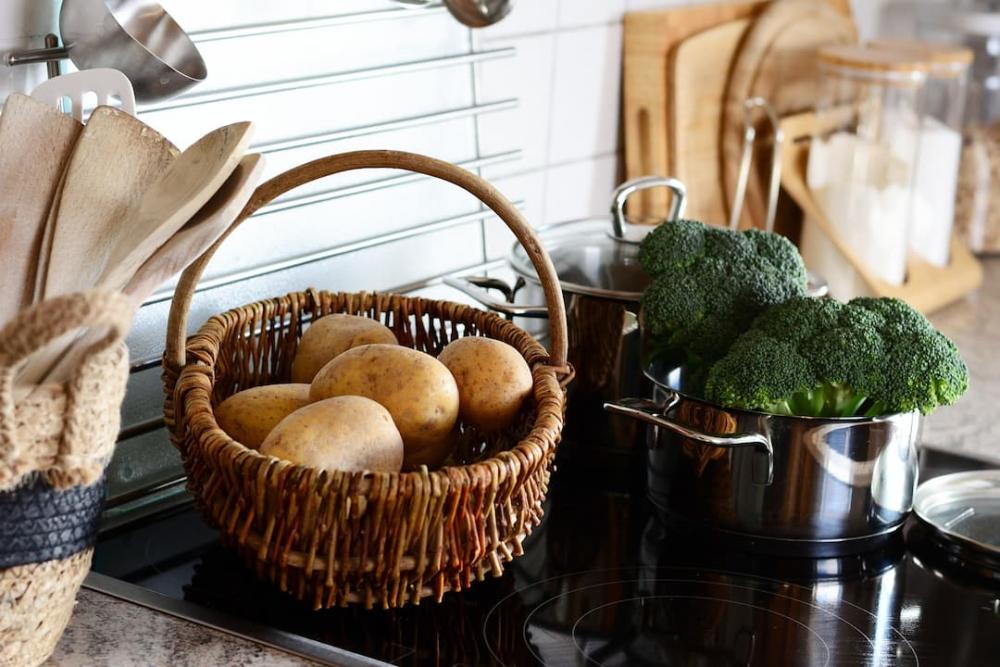 An induction hob with a basket of potatoes and pots of broccoli. There are glass containers, chopping boards, and a basket of spatulas nearby.