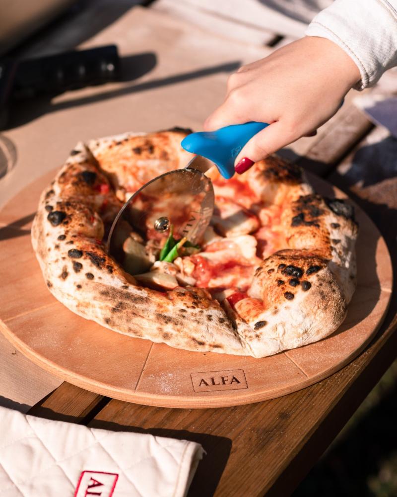 Homemade pizza with a crispy crust being cut using a cutter on an Alfa Forni wooden board.