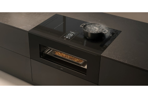 A modern induction hob and oven combo on a black island. There is a pot on the hob with boiling water, and the food in the oven is being baked.