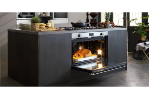 A kitchen island with a built-in stove and oven combo next to fruits, bread, plates, and a chopping board. A whole chicken with potatoes is seen in the open oven.