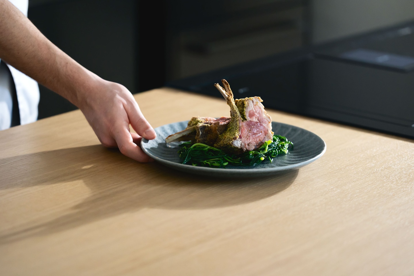 Chef placing a plate with herb-crusted rack of lamb on a bed of sautéed spinach, called Carré di Agnello, on a wooden table, conveying a gourmet dining atmosphere.