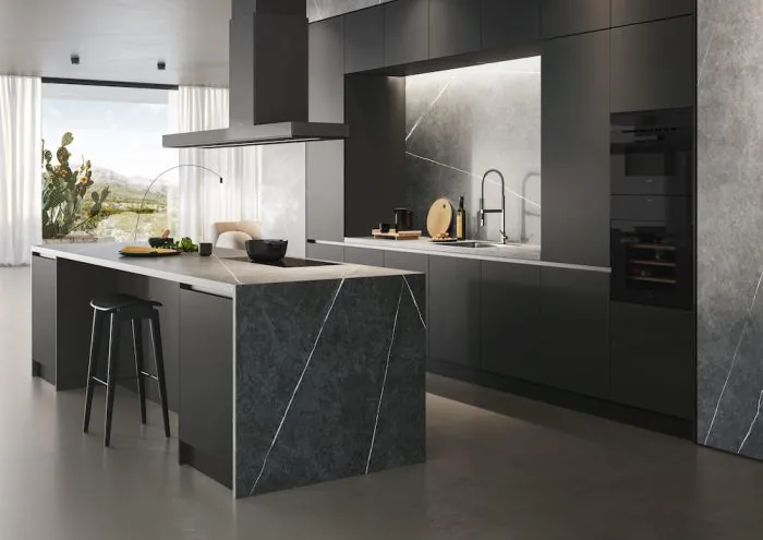 Modern kitchen with dark gray cabinets and an island featuring sleek, diagonal lines. Bright light streams through large windows illuminating a cactus.