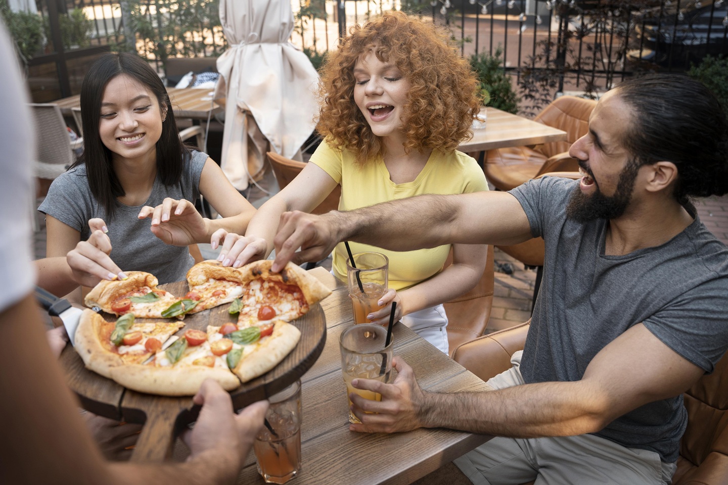 A group of three friends sits outdoors at a wooden table, smiling and sharing a pizza. They each hold drinks, creating a cheerful and relaxed atmosphere.