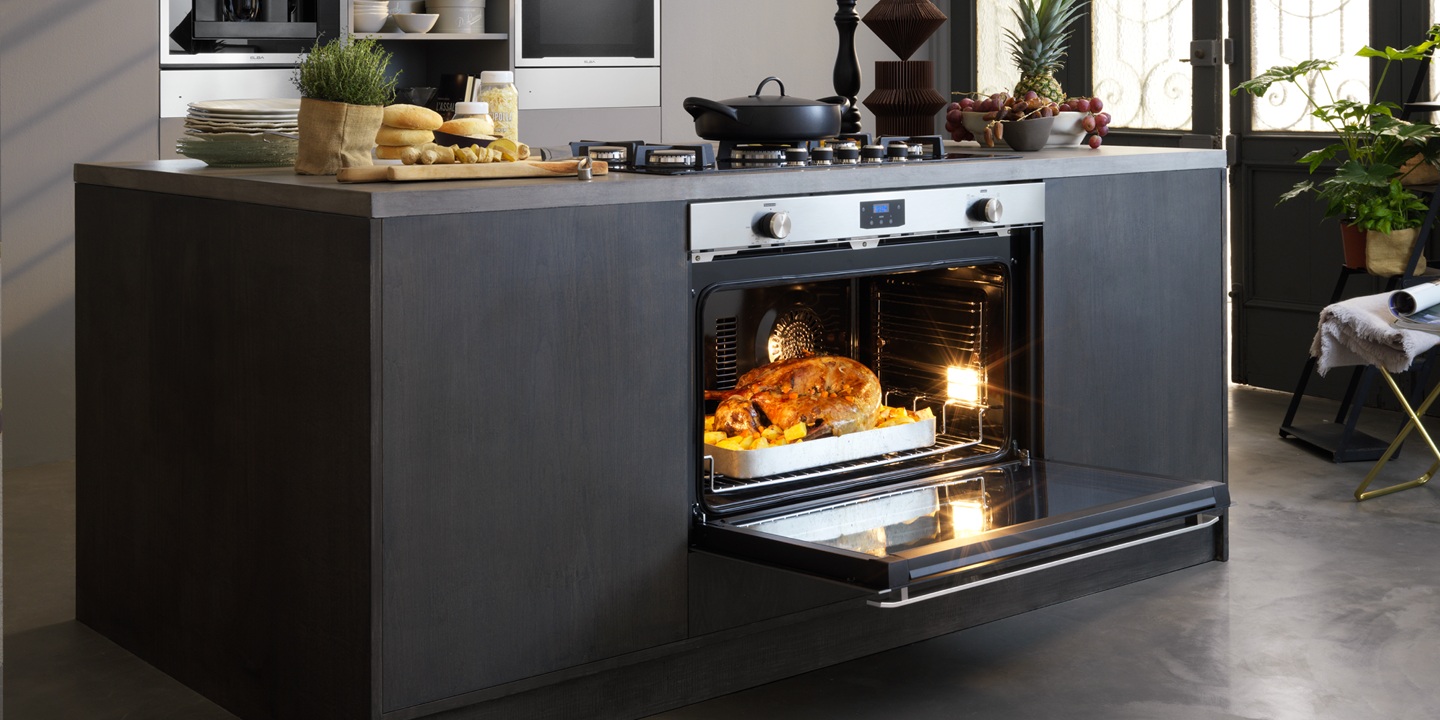 Modern kitchen with a central island featuring an open oven baking a golden-brown roast. Nearby are bread, fruit, and potted plants, creating a cozy ambiance. 