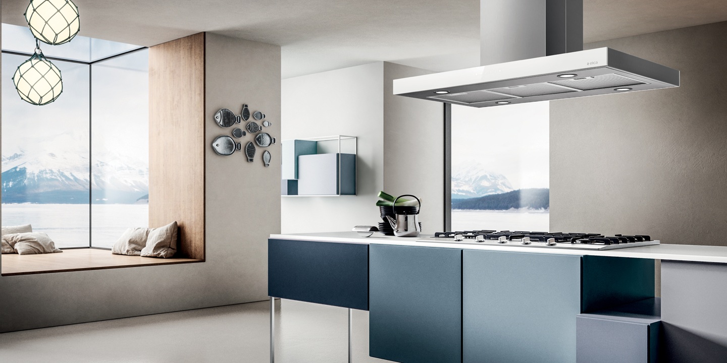 Modern kitchen with a sleek blue island and built-in stove under a range hood. An adjacent cozy nook features cushions and large windows with snowy mountain views. Minimalist decor and soft lighting create a calm ambiance. 
