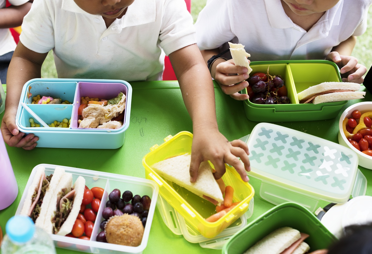 Children in white shirts enjoy lunch with colourful lunchboxes on a green table. Boxes contain sandwiches, fruits, and vegetables, evoking a playful, cheerful tone.