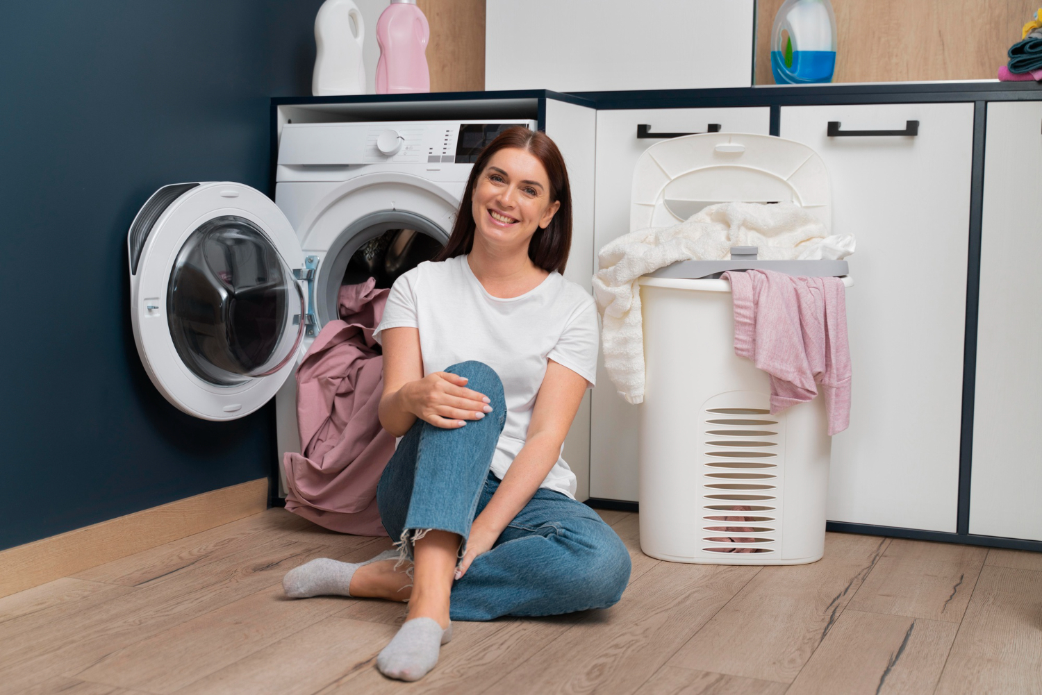 A woman is sitting on the floor in front of a washer and dryer combo with a basket of clothes beside her.
