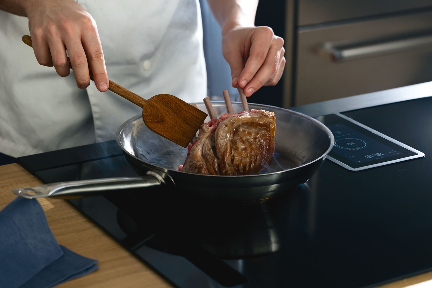 Chef in a white apron sears a rack of lamb in a silver pan on an Elica induction stove. The scene conveys a sense of focus and culinary precision.