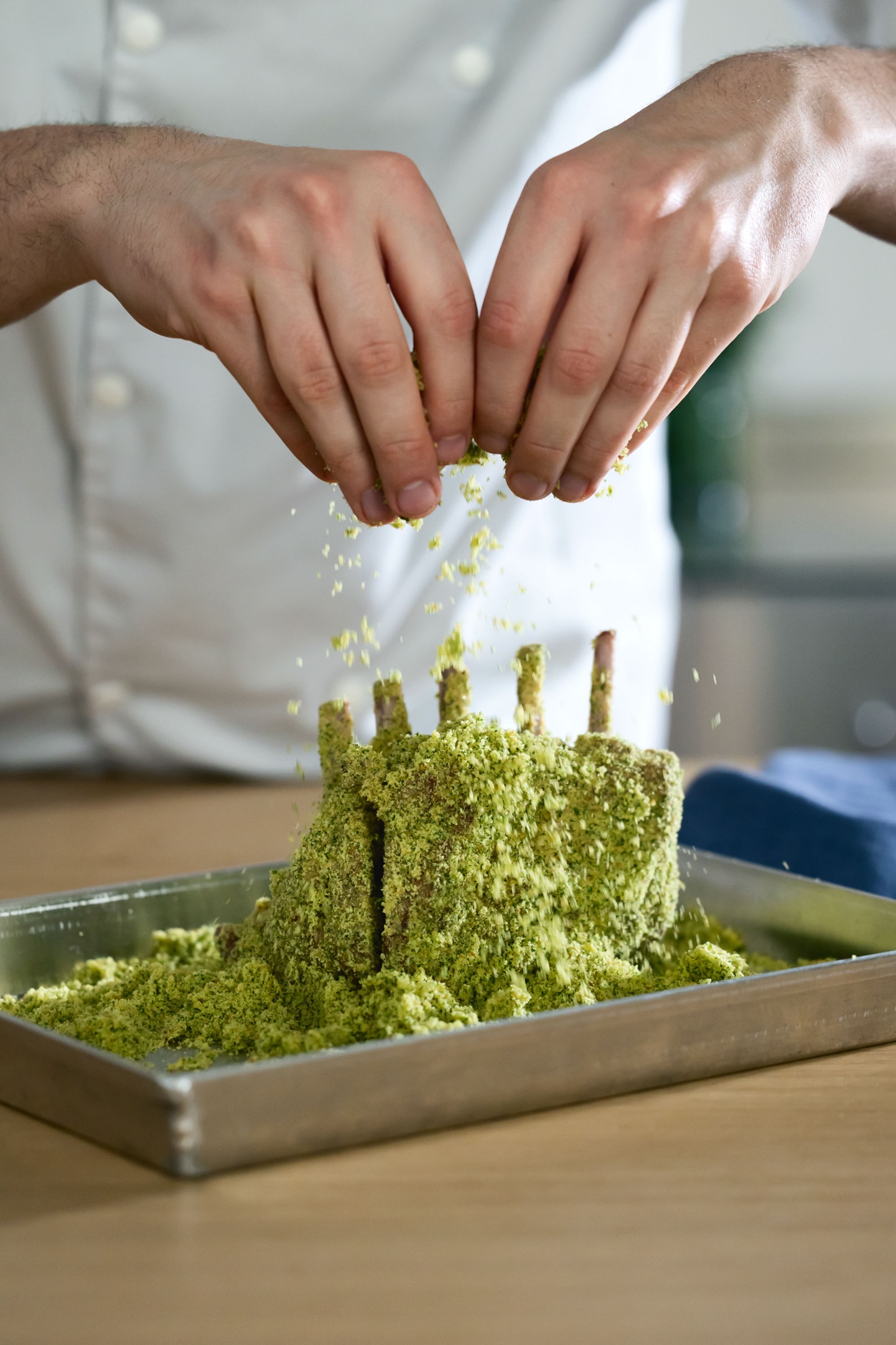 Hands sprinkle crushed pistachios onto a rack of lamb ribs standing in a metal tray. The background is a blurred kitchen setting, conveying a cooking scene.