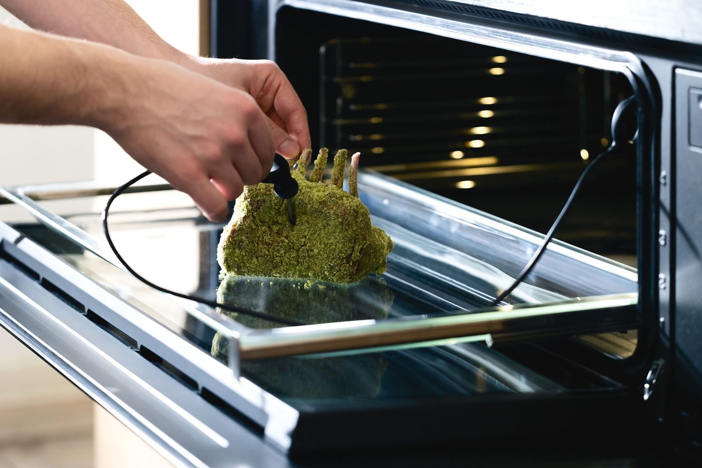 Chef places a meat thermometer into a herb-crusted rack of lamb inside an open oven, checking the temperature. The scene conveys careful cooking.