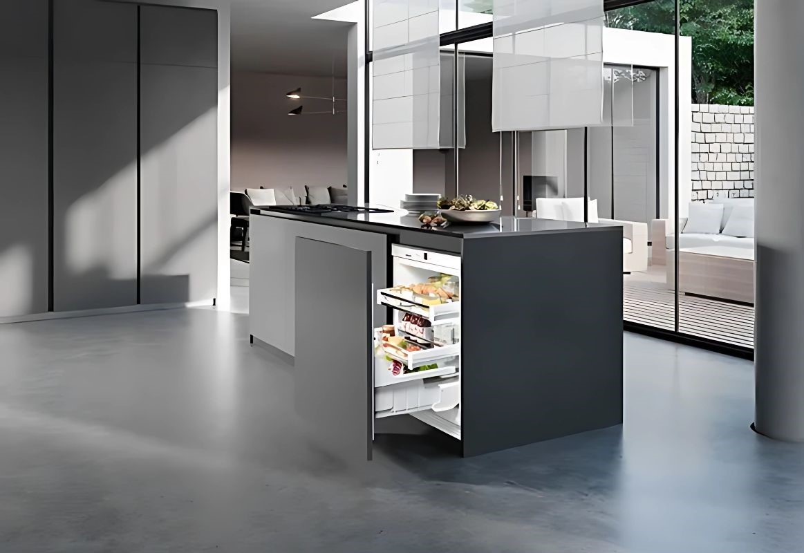 Sleek modern kitchen with a minimalist design. A central island features an open drawer fridge, stocked with food. Large windows and neutral tones give an airy feel.