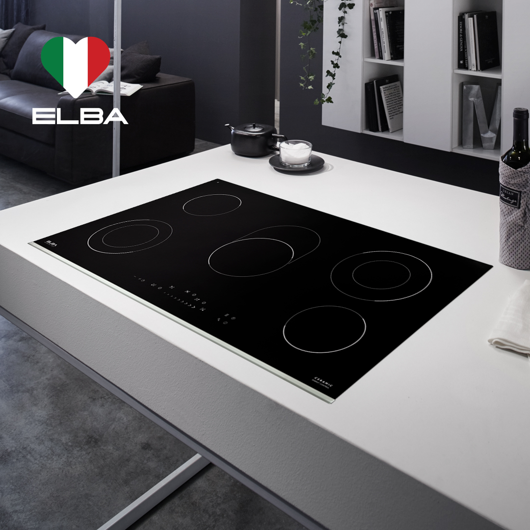 Modern kitchen with a sleek black induction cooktop on a white counter. Nearby are bookshelves, a pot, and a wine bottle, exuding a minimalist vibe.