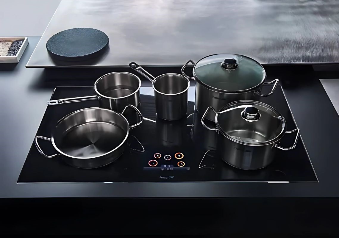 Five stainless steel pots and pans sit on a sleek black induction cooking hob with touch controls. The modern kitchen scene conveys elegance and functionality.