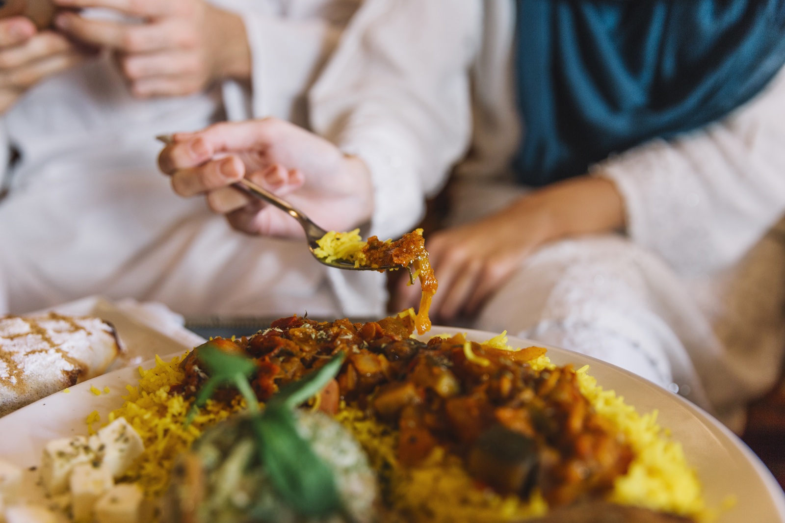 A hand holds a spoon of colorful stew above a plate of vibrant yellow rice. Two people in traditional attire sit together, creating a warm, communal feeling.