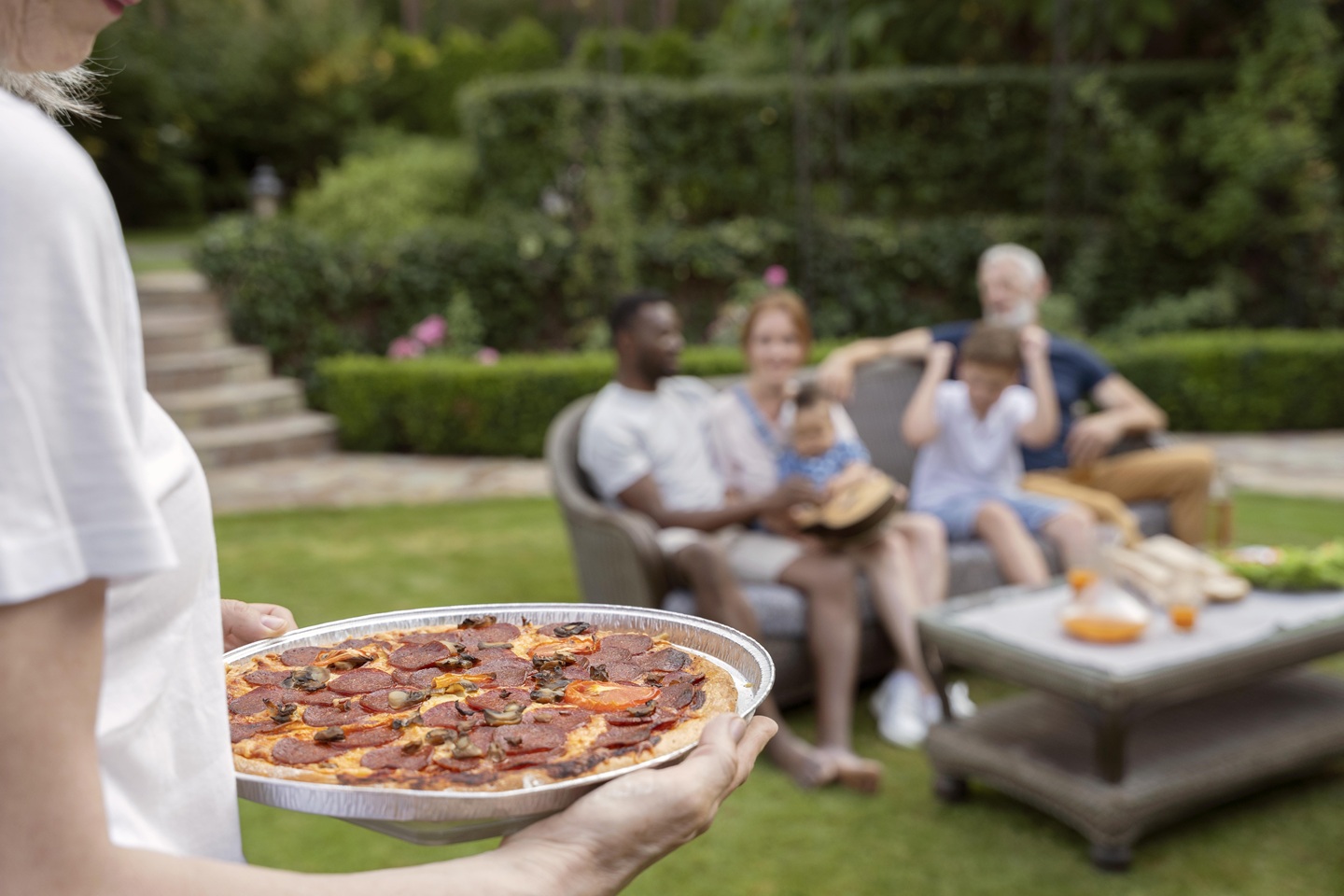 A person holds a tray of pepperoni pizza in the foreground, while a smiling family sits on outdoor furniture in a garden setting, conveying warmth and togetherness.