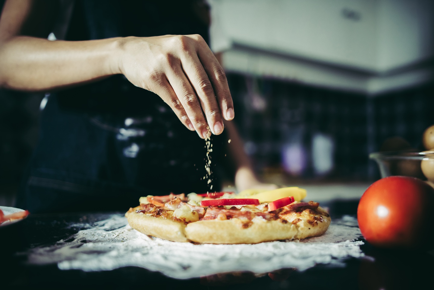A hand sprinkles cheese over a freshly baked pizza with sliced peppers. The kitchen scene conveys a warm, inviting atmosphere.