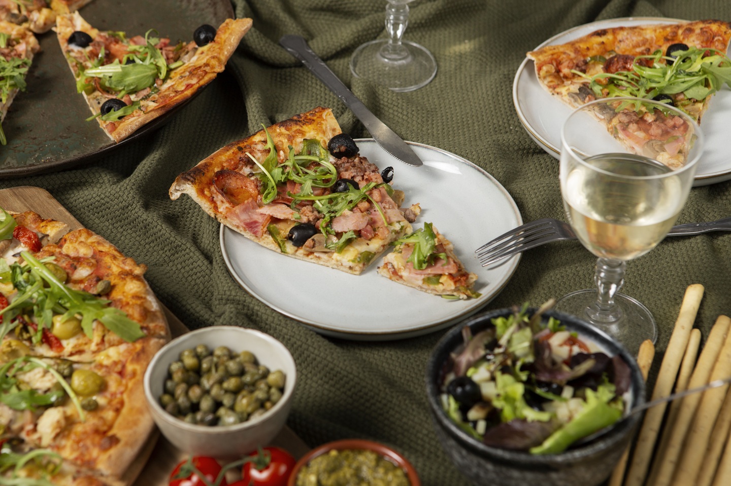 A cozy dining setup featuring pizzas topped with arugula, olives, and ham. A glass of white wine, salad, capers, and breadsticks complement the meal.