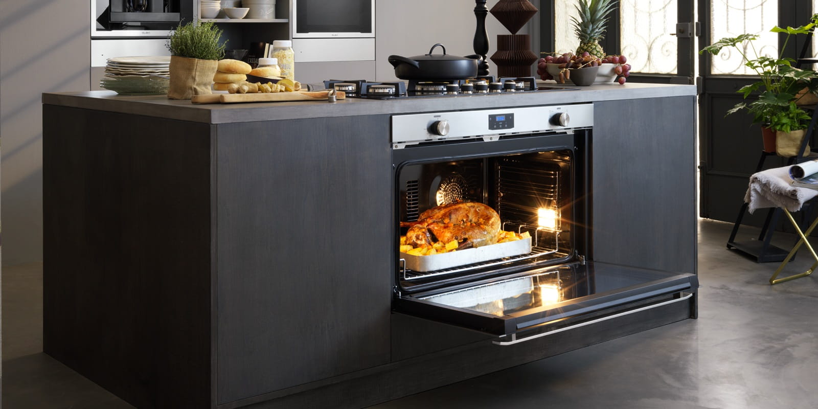 A modern kitchen with a central island features an oven cooking a golden-brown roast. The countertop is decorated with bread, herbs, and a potted plant, evoking a cozy, inviting atmosphere.