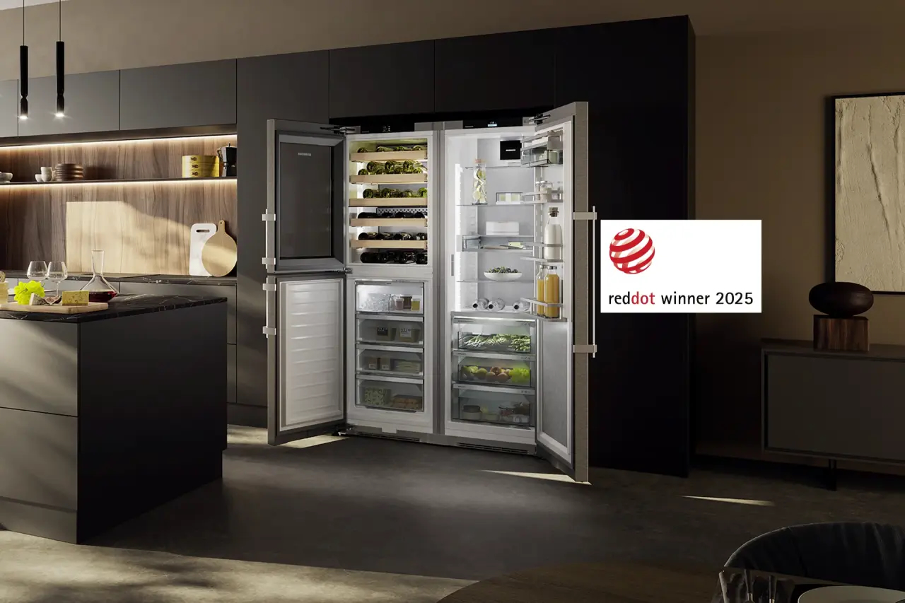 Modern kitchen with an open double-door Liebherr refrigerator filled with organised food and beverages, showcasing a Red Dot Award 2025 logo. The ambiance is sleek and sophisticated.