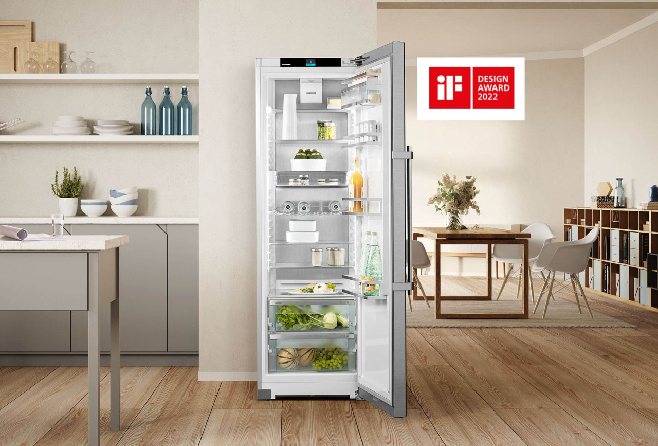 Modern kitchen with an open stainless steel fridge by Liebherr stocked neatly with food. Minimalistic decor, wooden floor, and iF Design Award 2022 plaque.