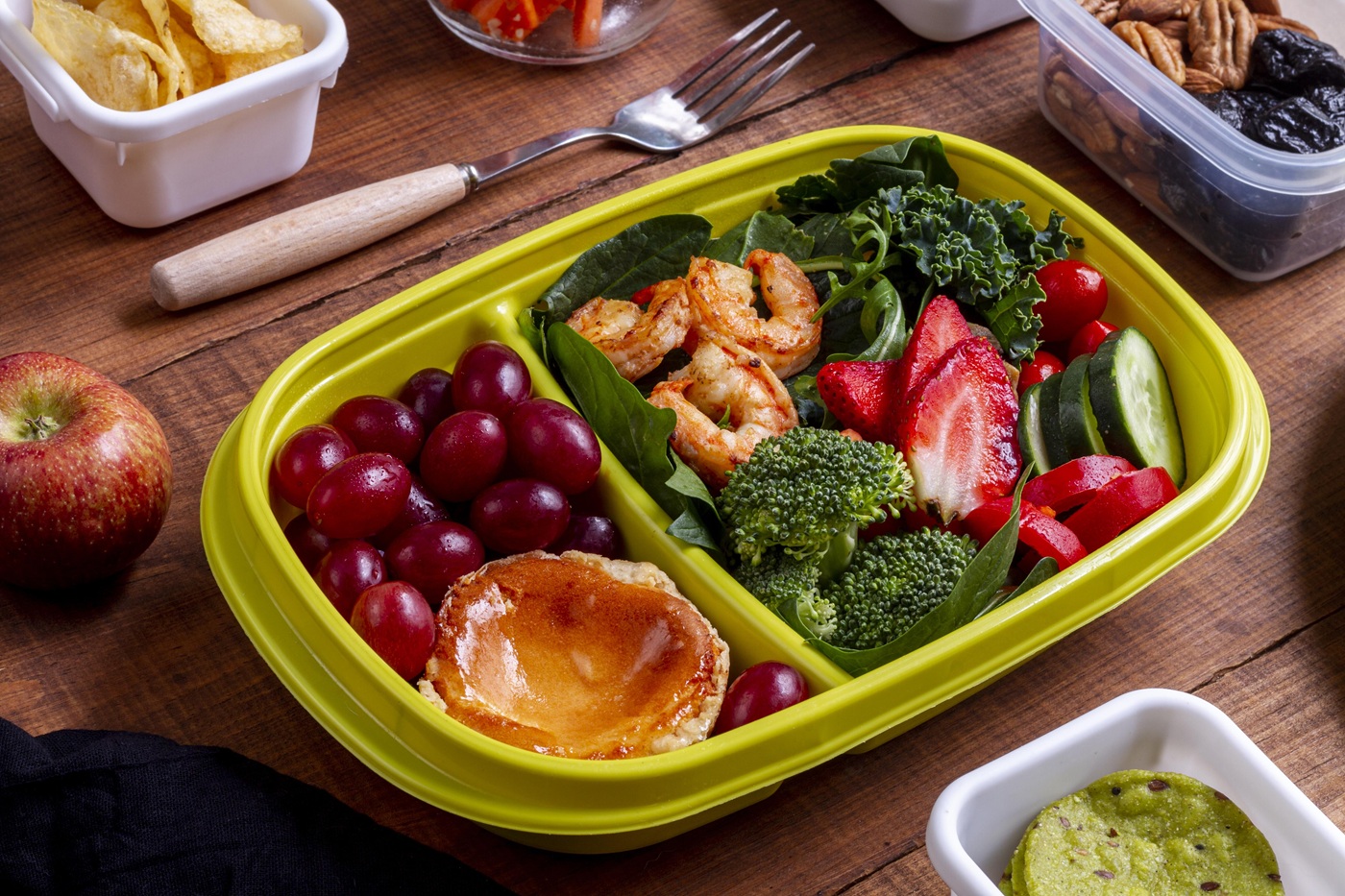 A lunchbox meal in a yellow tray with spicy shrimp, fresh greens, strawberries, broccoli, grapes, and a small pastry on a wooden table. Nearby are an apple, chips, nuts, and dip.