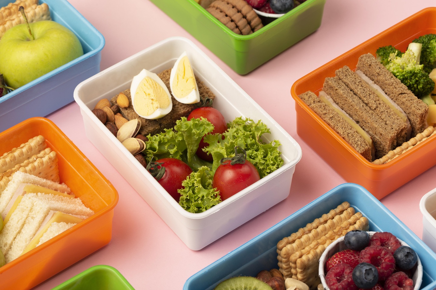 Colourful lunchboxes on a pink background contain sandwiches, fruits, vegetables, nuts, and eggs, presenting a fresh, healthy meal variety.