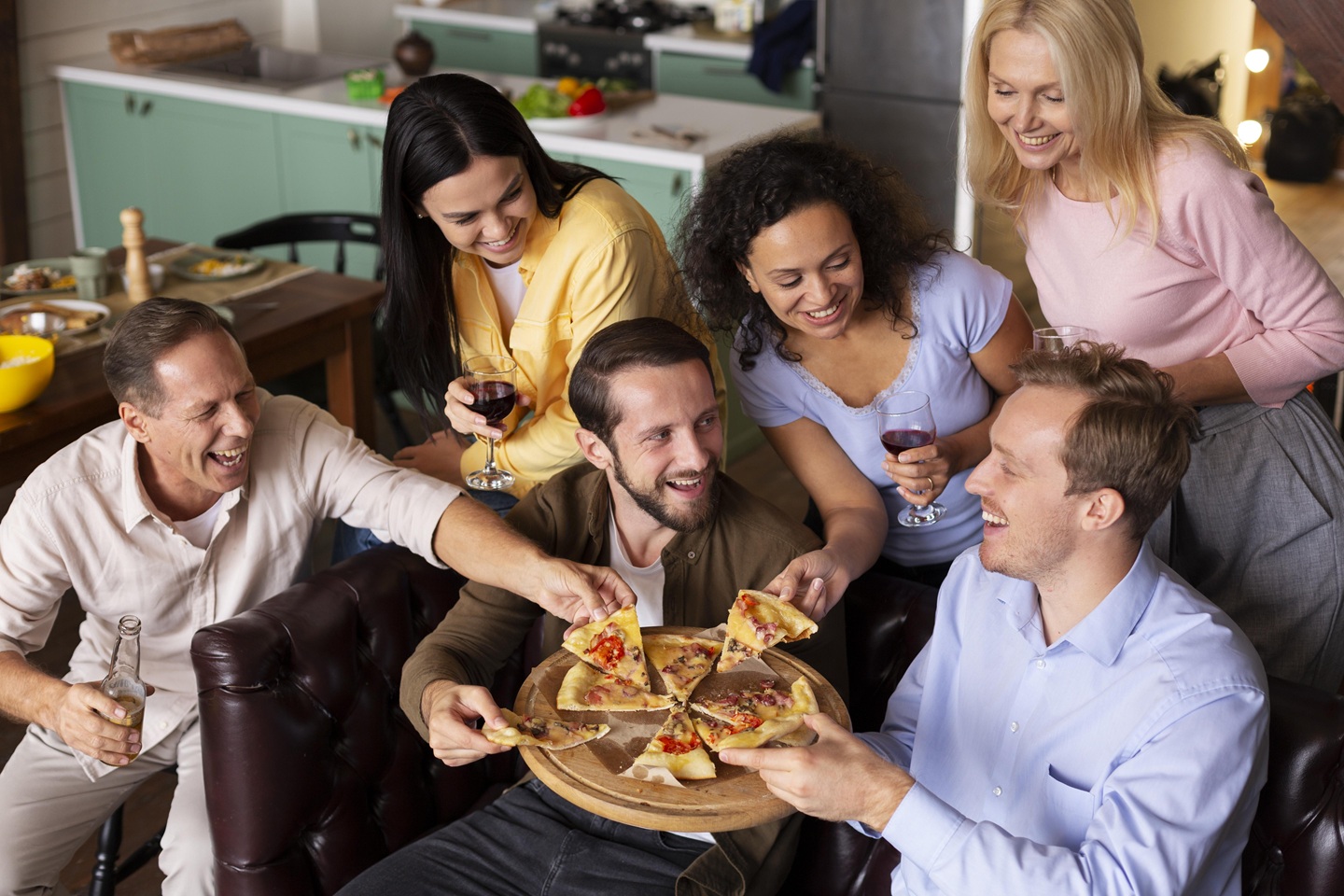 Group of six friends happily sharing pizza in a cozy kitchen, holding drinks. They are smiling and engaged, conveying a joyful and warm atmosphere.