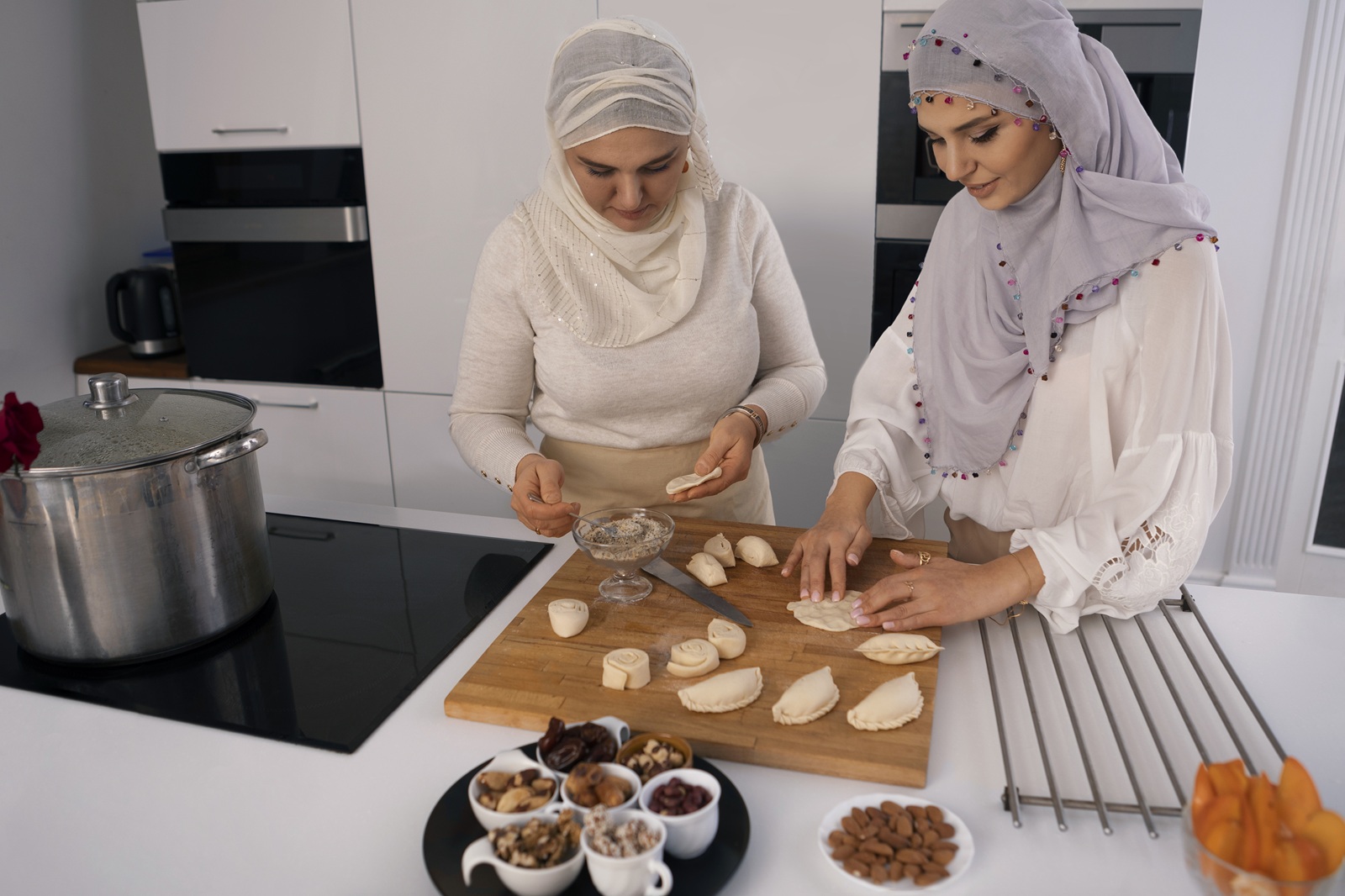 Two women wearing headscarves are in a modern kitchen making pastries. They focus intently on filling dough, surrounded by small bowls of nuts and dates.